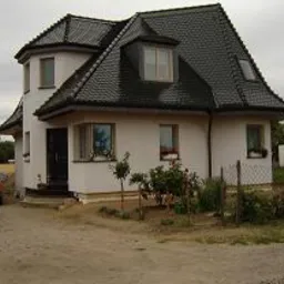 Nietypowy, parterowy dom z poddaszem użytkowym, o zaokrąglonych narożnikach i ciemnym dachu z dachówki ceramicznej, widoczny z perspektywy ogrodu, z żółtym samochodem zaparkowanym obok.